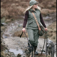 Woman in hunting attire with a green vest and cap, holding a rifle, in a natural setting.